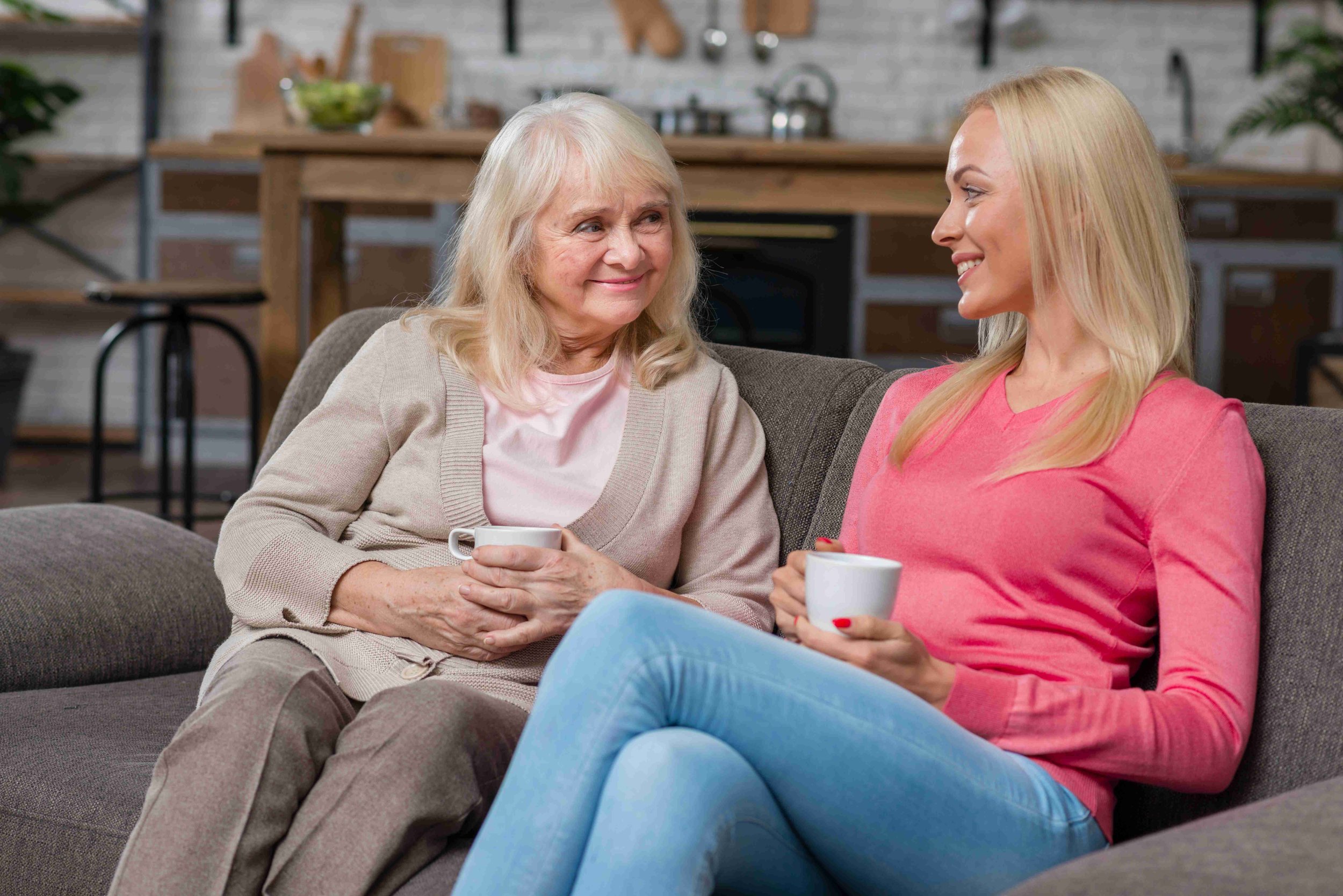 Two women sitting together on a couch with coffee, laughing and talking easily