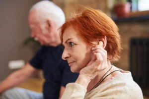 An elderly woman with red hair adjusting a hearing aid in her ear, sitting indoors with a blurred elderly man in the background - Adirondack Audiology